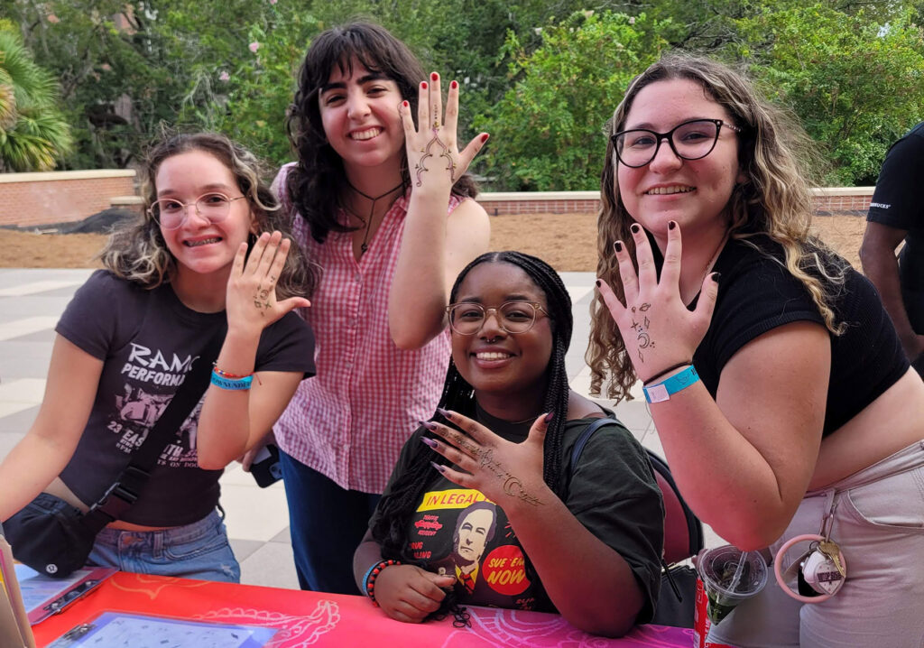 Picture of 4 young women holding up their hands to show off their henna designs with big smiles. Picture is outside.