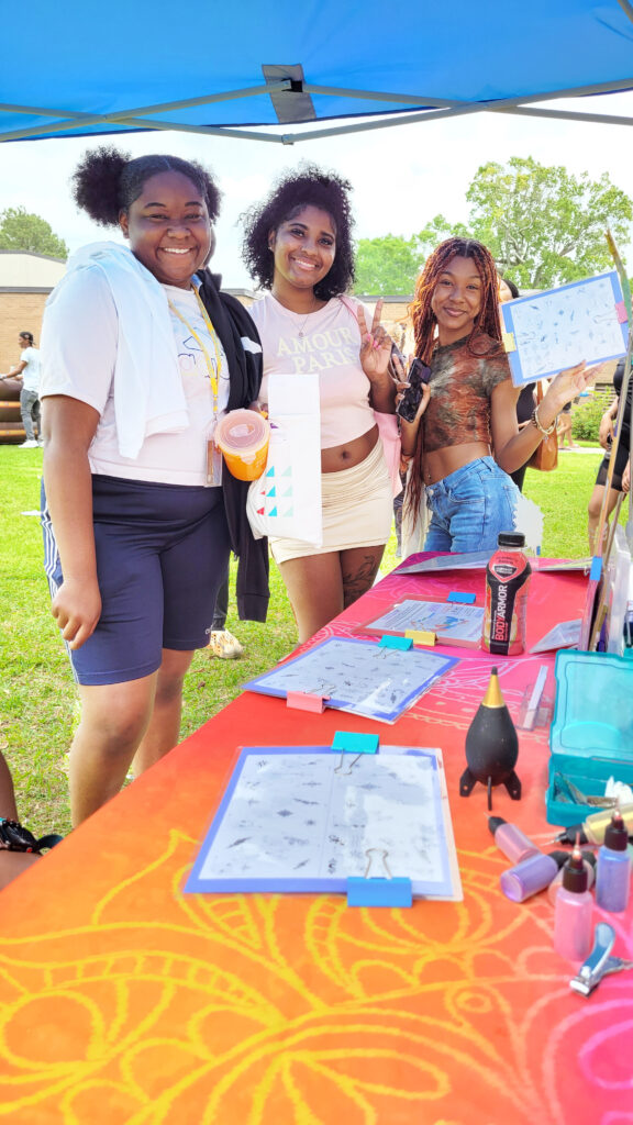 Three students in line at The Henna Experience Tent with big smiles and the table in the foreground.
